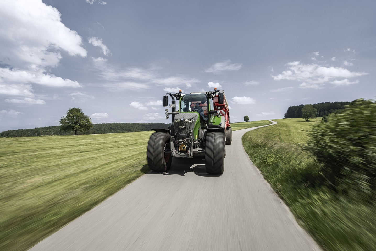 Ein grüner Fendt 700 Vario Gen7.1 fährt auf einer Landstraße im Sommer zwischen grünen Feldern links und rechts