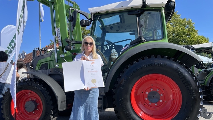 Eine Frau mit einer weißen Bluse und einem Jeansrock hält eine Urkunde mit einer Goldmedaille, im Hintergrund der grüne Fendt e100 Vario Traktor