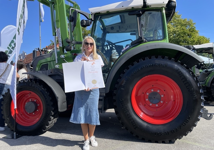 Alja Curkovic (Marketingleiterin Inter Agrar) mit dem Award vor dem Fendt e100 Vario Eine Frau mit einer weißen Bluse und einem Jeansrock hält eine Urkunde mit einer Goldmedaille, im Hintergrund der grüne Fendt e100 Vario Traktor