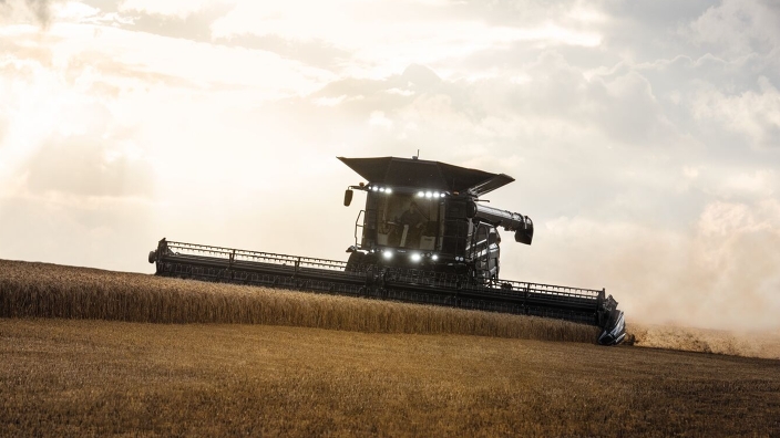 Ein Fendt IDEAL Mähdrescher fährt frontal auf die Kamera zu und drischt Weizen im frühen Abend. Die Scheinwerfer sind angeschaltet.
