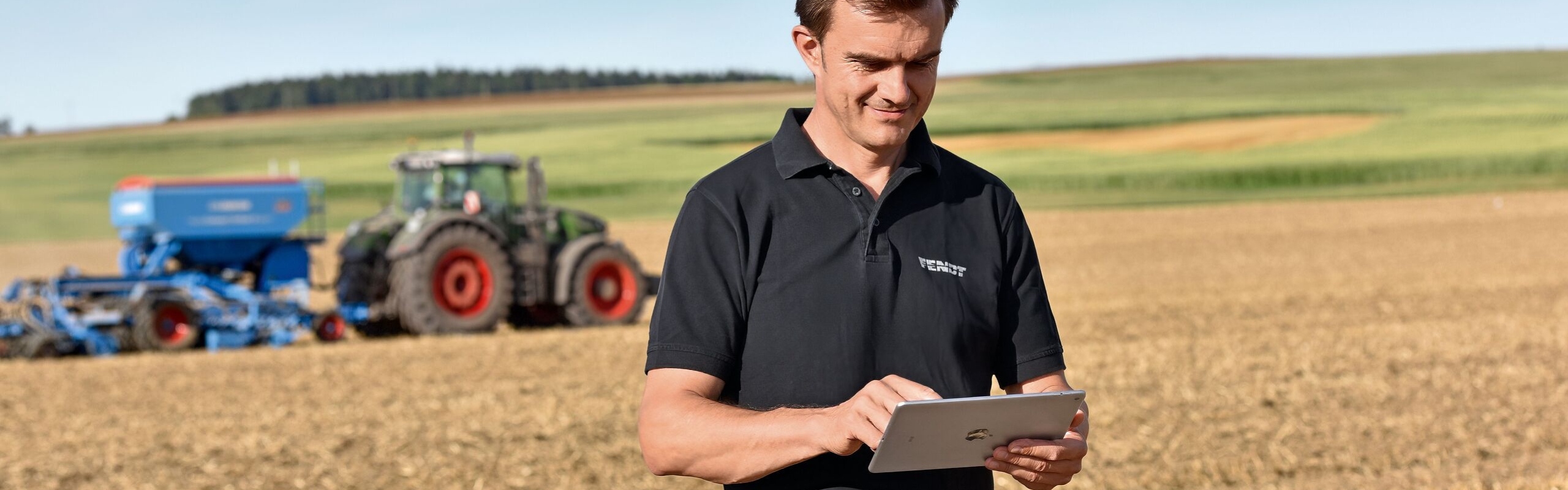 Ein Landwirt auf dem Feld mit Tablet in der Hand, im Hintergrund sieht man seinen Fendt Traktor mit Anbaugerät