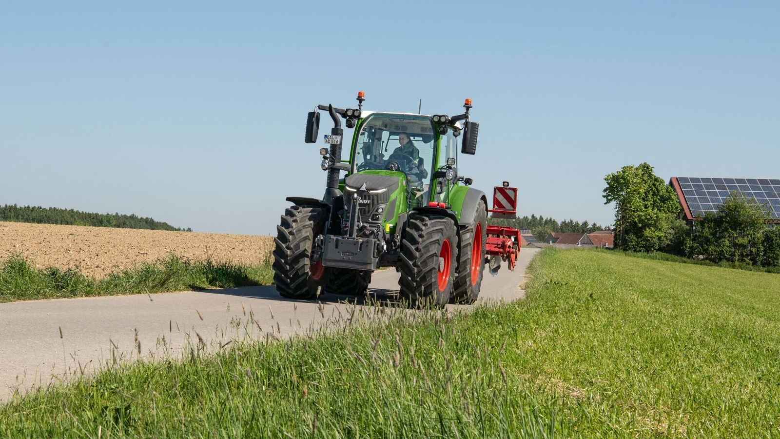 Fendt 500 Vario Traktor auf der Straße Ein Fendt 500 Vario Gen4 in grün mit roten Felgen fährt auf einer Straße. Links von der Straße ist ein Acker und rechts davon ein grüne Wiese. Auf der Wiese sieht man im Hintergrund Häuser.