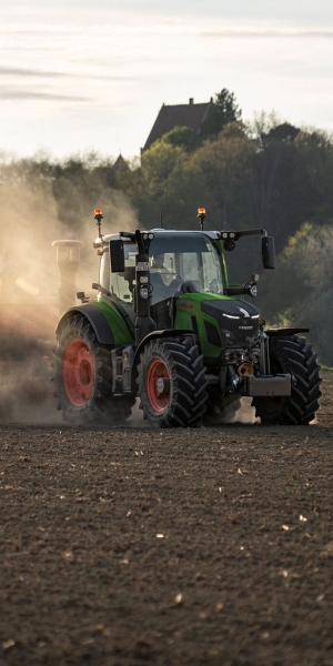 Fendt 500 Vario Traktor auf dem Acker Ein Fendt 500 Vario Gen4 Traktor in grün mit roten Felgen fährt auf einer hellgrünen Wiese. Hinten ist ein rotes Bodenbearbeitungsgerät mit Warntafel angehängt. Im Hintergrund sind Felder, ein Waldrand und blauer Himmel mit wenigen Wolken zu sehen.