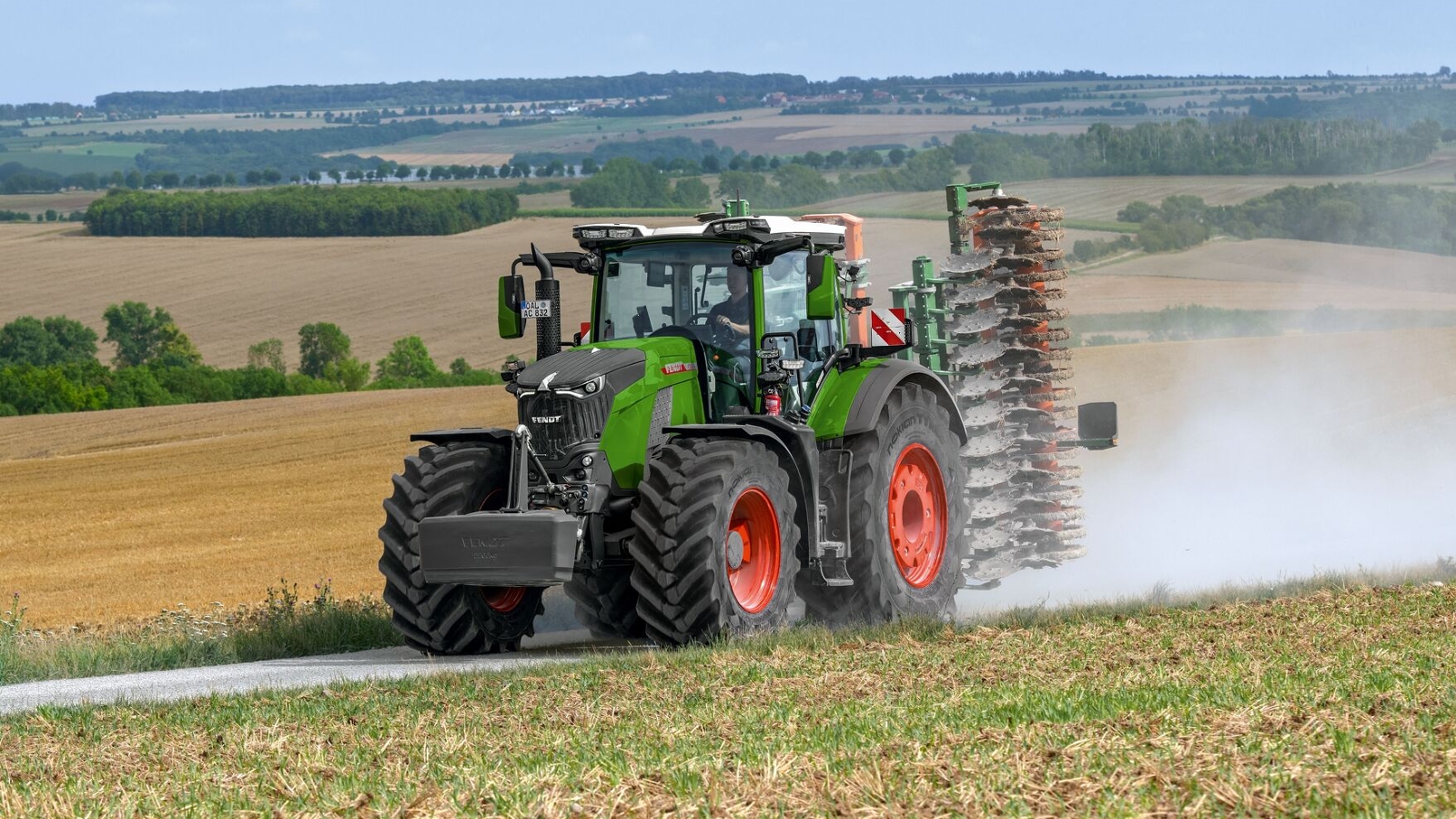 Fendt 800 Vario Gen5 auf einem Feldweg Der Fendt 800 Vario Gen5 in grün mit roten Felgen und im Heck mit einer klappbaren Scheibenegge auf einem Kiesweg. Im Hintergrund sind Felder und Bäume zu sehen.