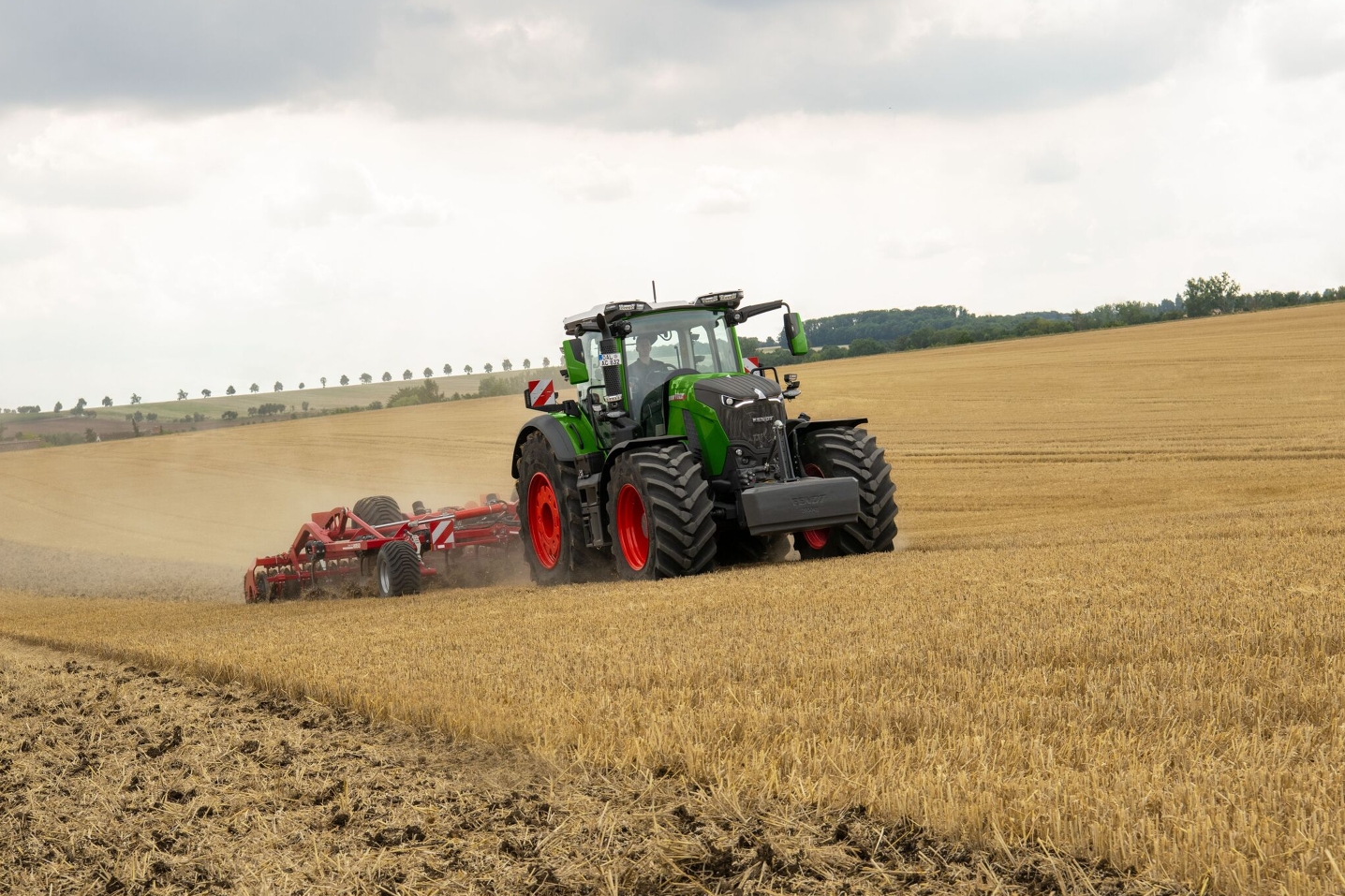 Fendt 800 Vario Gen5 auf einem Feld mit Anbaugerät Der Fendt 800 Vario Gen5 in grün mit rotem Felgen fährt auf einem Feld mit Strohstoppeln. Man sieht, dass er im Heck ein rotes Anbaugerät hat, was ein Gruppe ist. Im Hintergrund sieht man Bäume und den Himmel.