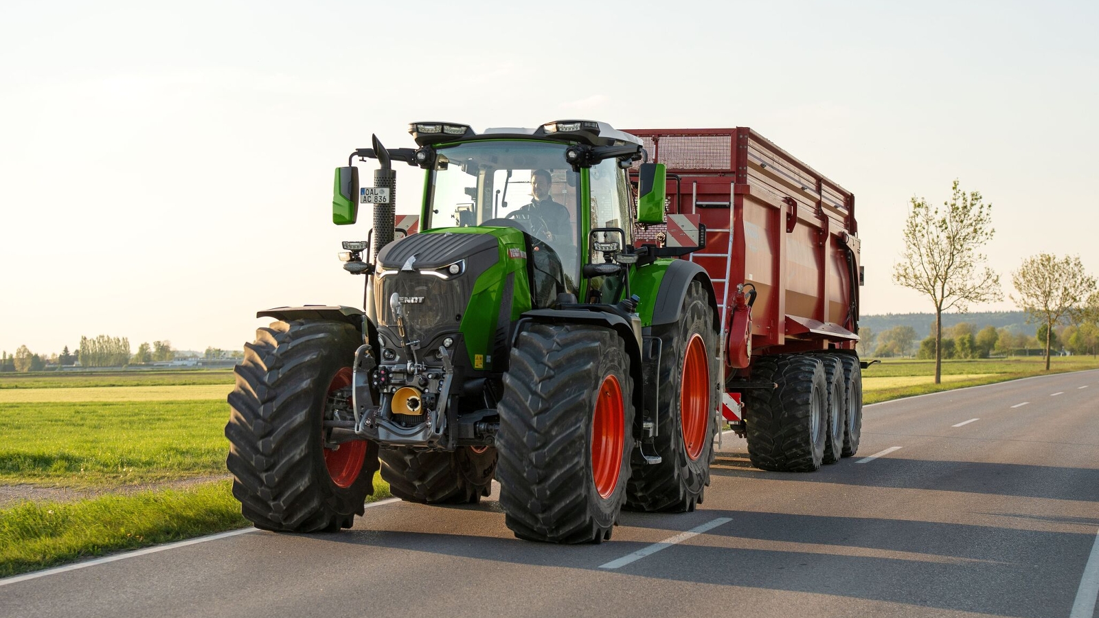 Ein grüner Fendt 800 Vario Gen5 mit roten Felgen fährt auf der Straße und hat im Heck einen roten Ladewagen, den er zieht. Links neben der Straße ist eine grüne Wiese und im Hintergrund sieht man vereinzelt Bäume.