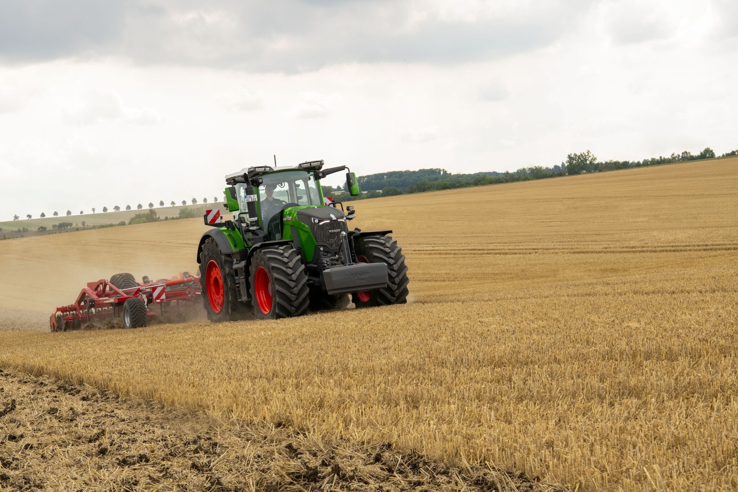 Ein Fendt 800 Vario Gen5 mit roten Felgen fährt über ein Feld und hat im Heck einen roten Grubber zur Bodenbearbeitung angehängt. Im Hintergrund sieht man vereinzelt Bäume und rechts hinten im Bild einen Wald