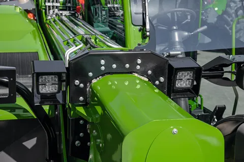 Forfra-billede af en grøn Fendt Cargo T740 frontlæsser. I forgrunden ses den massive, grønne bomarm med sort fastgørelsesplade og to rektangulære LED-arbejdslygter. Bagved ses førerkabinen med rat og betjeningselementer samt de hydrauliske ledninger, der