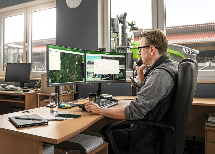 Mand sidder ved skrivebord foran FendtONE webapplikation og Fendt traktorer i baggrunden
