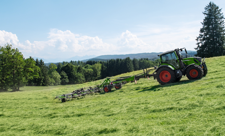 En grøn Fendt traktor med røde fælge og det grønne Fendt Lotus redskab i arbejde med græshøstning på en grøn eng med blå himmel og udsigt over landskabet.