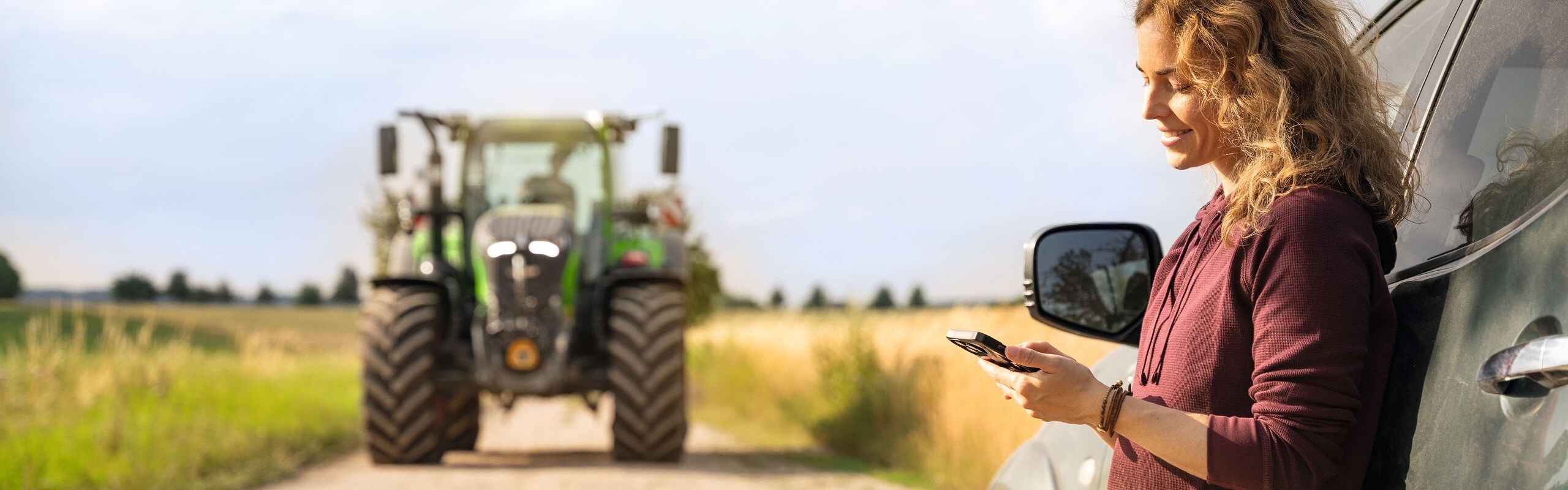 En kvinde med lyse krøller, der bærer en mørkerød trøje står og læner sig op ad en bil mens hun ser på sin mobil, i baggrunden kan man se en Fendt-traktor