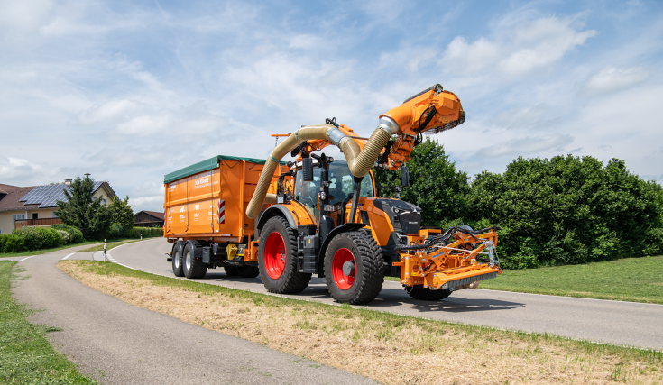 Fendt 728 Vario på landevejen med en slåmaskine En kommunal orange Fendt 728 Vario kører med en bomklipper og en trailer på en vej i transport, med en landsby i baggrunden.