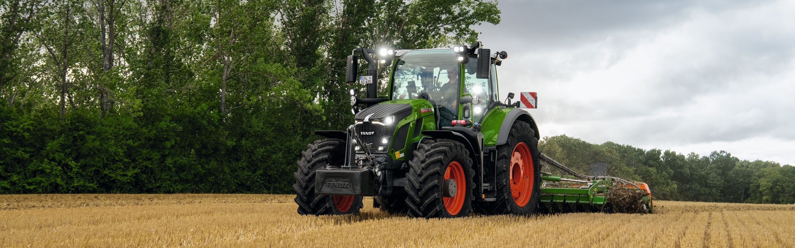 Den grønne Fendt 600 Vario traktor med røde fælge på arbejde på en mark med træer i baggrunden og en skyet himmel.