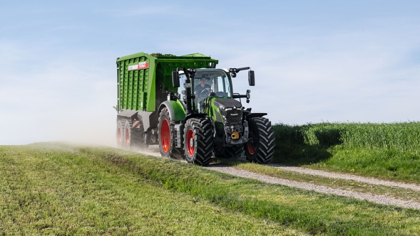 Den grønne Fendt 600 Vario traktor med røde fælge og den grønne Fendt læssevogn Fendt Tigo kører på en lille markvej med en grøn eng i for- og baggrunden.