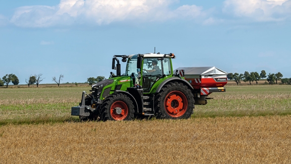 Den grønne traktor Fendt 600 Vario med røde fælge kører set fra siden på en grøn eng. I baggrunden ses den blå himmel med hvide skyer.