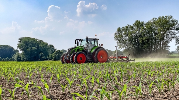 Den grønne traktor Fendt 600 Vario med tvillingedæk på arbejde på en mark med afgrøder. I baggrunden ses mørkegrønne træer og en lyseblå himmel med hvide skyer,