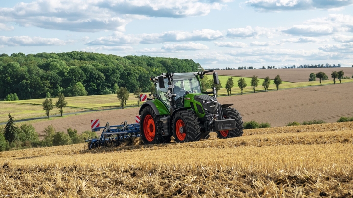 En grøn Fendt traktor med røde fælge ved jordbearbejdning på en høstet mark med et blødt bakkelandskab i baggrunden