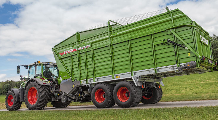 Grøn landbrugsvogn med påskriften »Fendt Tigo XR«, koblet til en traktor. Vognen er udstyret med store røde hjul og står på en eng under en klar himmel