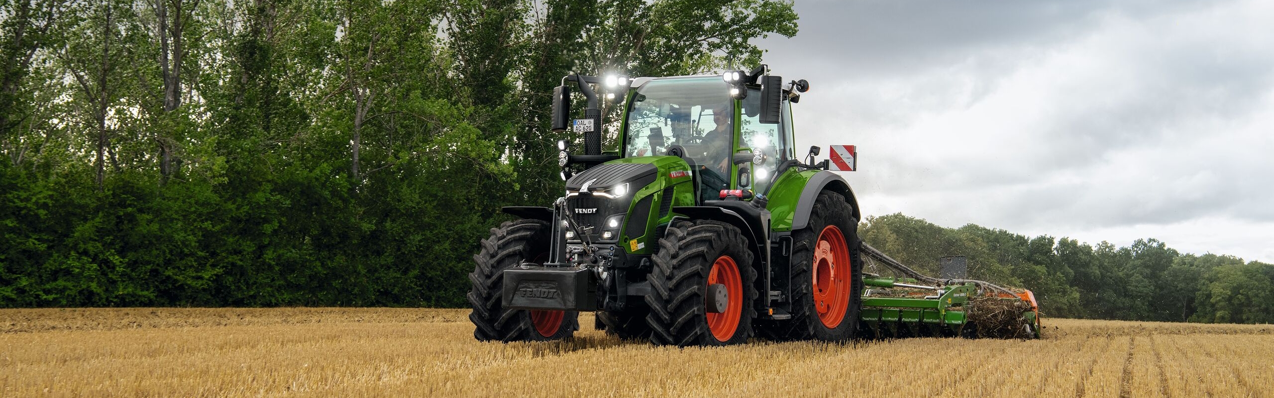 Den grønne Fendt 600 Vario-traktor med røde fælge i brug på en mark med træer i baggrunden og en overskyet himmel.