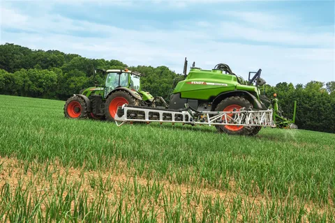 Un tractor Fendt verde con un pulverizador agrícola Fendt Rogator 366 verde pulveriza productos fitosanitarios sobre un prado verde, con un bosque al fondo.