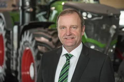 Retrato de Andreas Loewel con camisa blanca, chaqueta negra y corbata a rayas verdes, con un gran tractor Fendt verde al fondo.