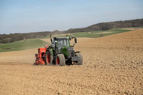 Un Fendt 300 Vario verde con una sembradora roja y un contrapeso delantero circula por un campo durante la siembra.