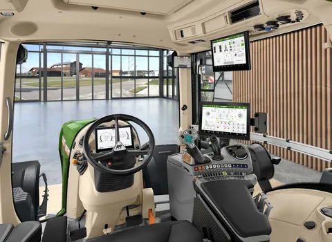 Vista interior de la cabina del tractor desde la perspectiva del conductor con volante, reposabrazos de control FendtONE con joystick y terminal, así como un terminal adicional en el techo. A través de la ventana delantera se ve un vestíbulo con una gran
