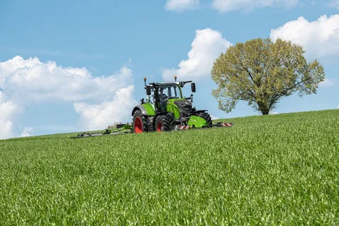 Un Fendt 700 Vario Gen7 verde siega un prado con una segadora delantera y trasera, al fondo se ve un árbol.