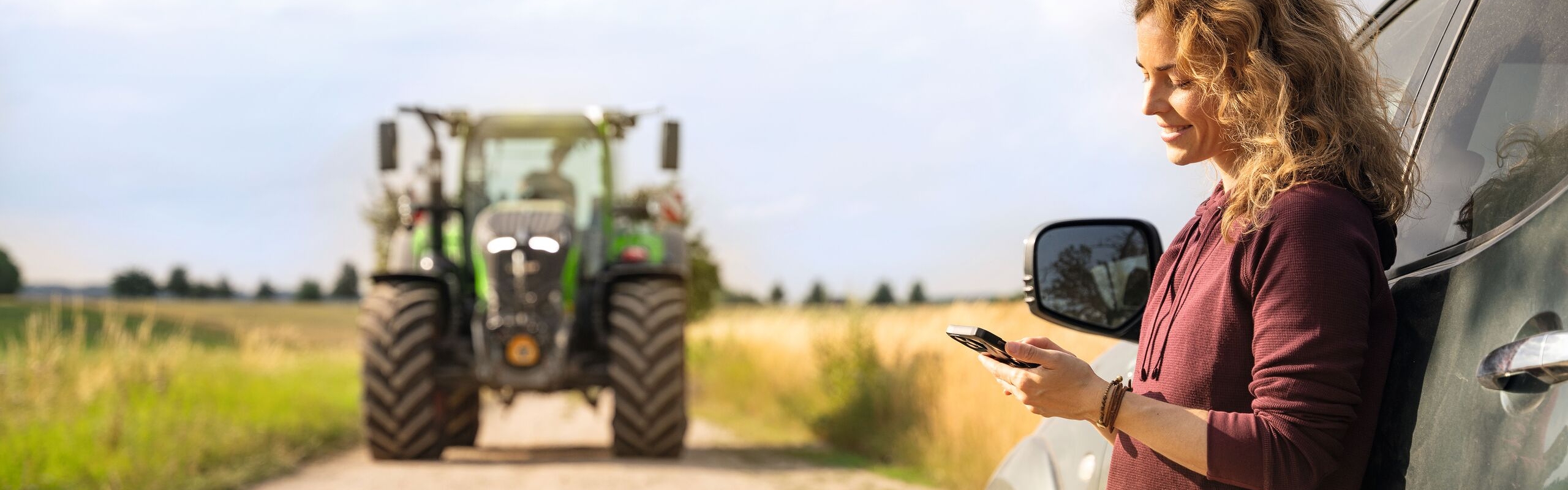 Una mujer con una melena rubia, rizada y un jersey rojo está apoyada en su automóvil mirando al móvil; al fondo se ve un tractor Fendt.