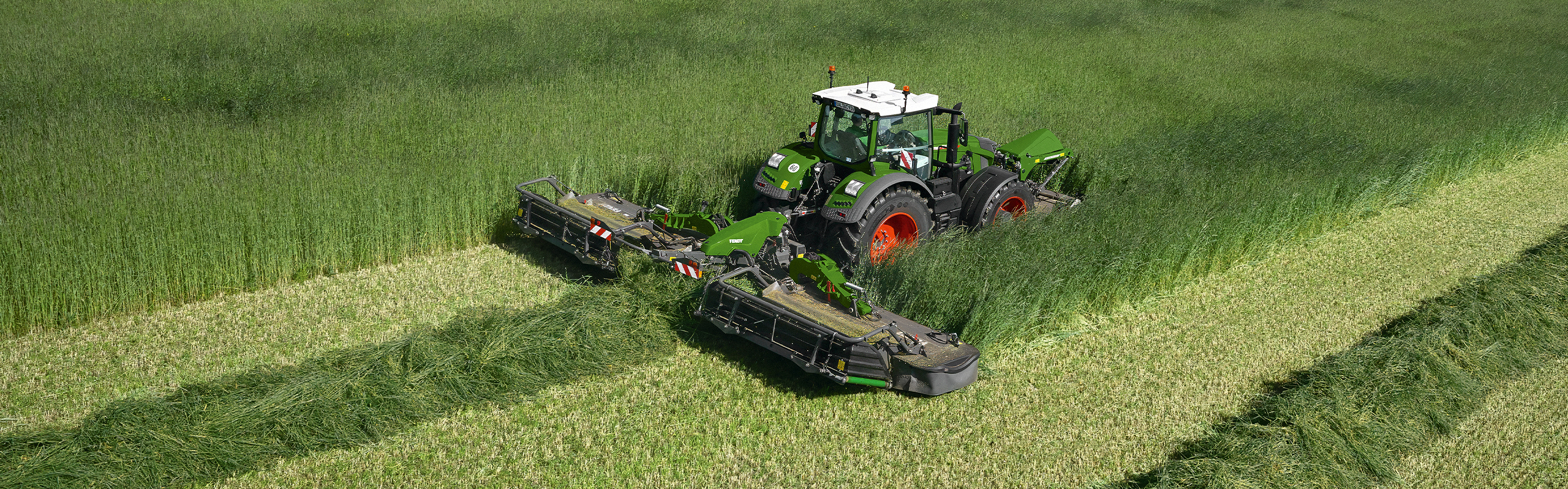 Un agricultor siega con una segadora Fendt Slicer frontal y trasera por prado verde.