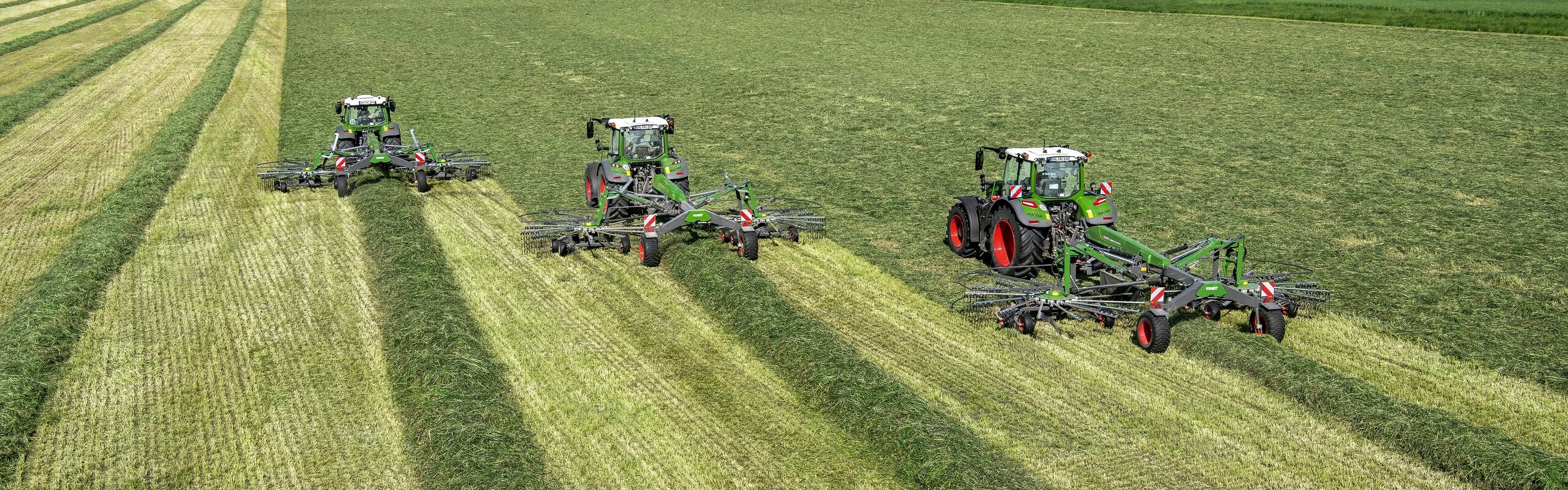 Tres tractores Fendt verdes con rastrillos hileradores Fendt Former montados, mientras trabajan un campo bajo el cielo azul