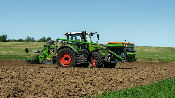 El tractor Fendt 600 Vario verde con llantas rojas y una trilladora como implemento circula sobre una parcela. Al fondo se ven un prado verde y un cielo azul.