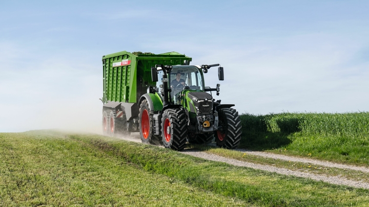El tractor Fendt 600 Vario verde con llantas rojas y el remolque autocargador Fendt Tigo circula por una pista asfaltada en medio de prados verdes.