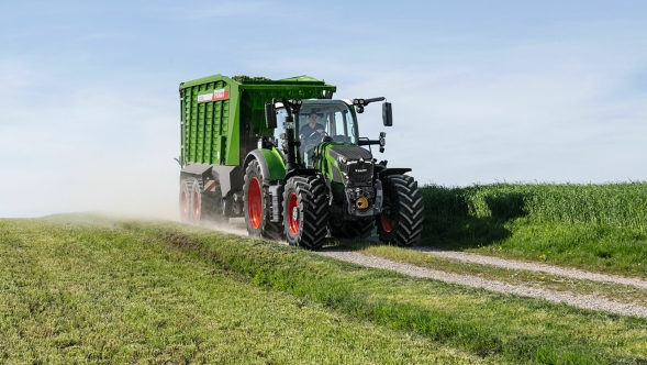 El tractor Fendt 600 Vario verde con llantas rojas y el remolque autocargador Fendt Tigo circula por una pista asfaltada en medio de prados verdes.