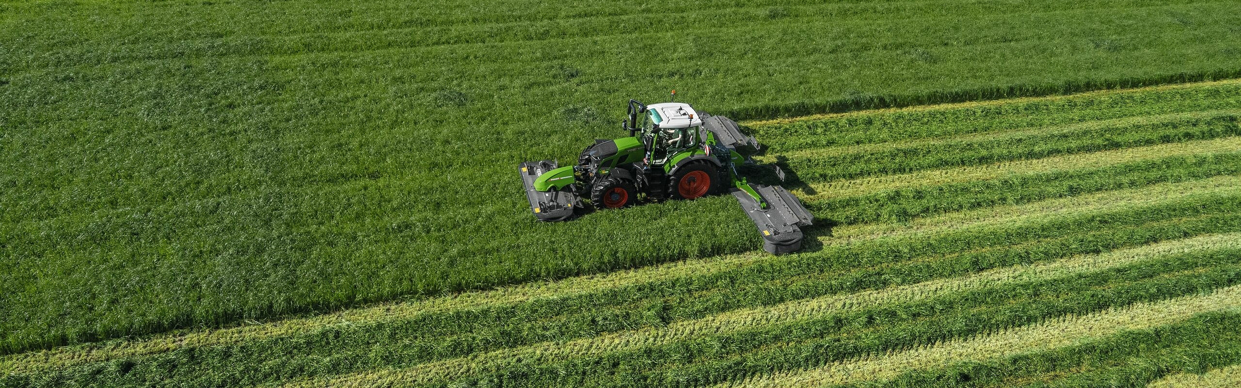 Vista de pájaro del tractor verde Fendt 600 Vario trabajando con una segadora Fendt Slicer en un gran prado verde.