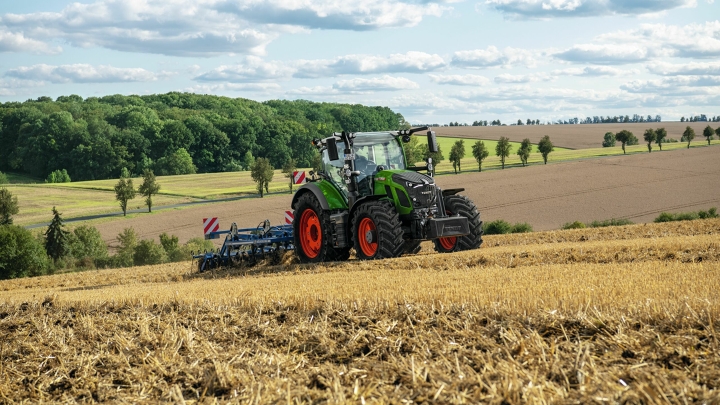 Un tractor Fendt verde con llantas rojas trabajando el suelo en un campo cosechado con un paisaje de montículos al fondo.