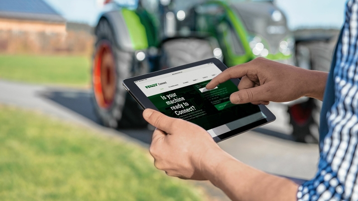 Primer plano de una tableta en la mano de un agricultor que verifica sus datos centrales de máquina a través de Fendt Connect. Al fondo, se ve ligeramente borroso un tractor verde de la marca Fendt.