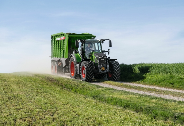 Fendt 600 Vario El tractor Fendt 600 Vario verde con llantas rojas y el remolque autocargador Fendt Tigo circula por una pista asfaltada en medio de prados verdes.