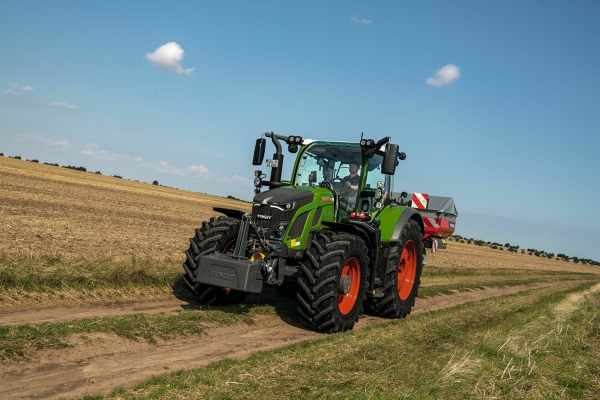 Fendt 600 Vario El tractor Fendt 600 Vario verde con llantas rojas circula por una pequeña pista asfaltada con un cielo celeste al fondo.