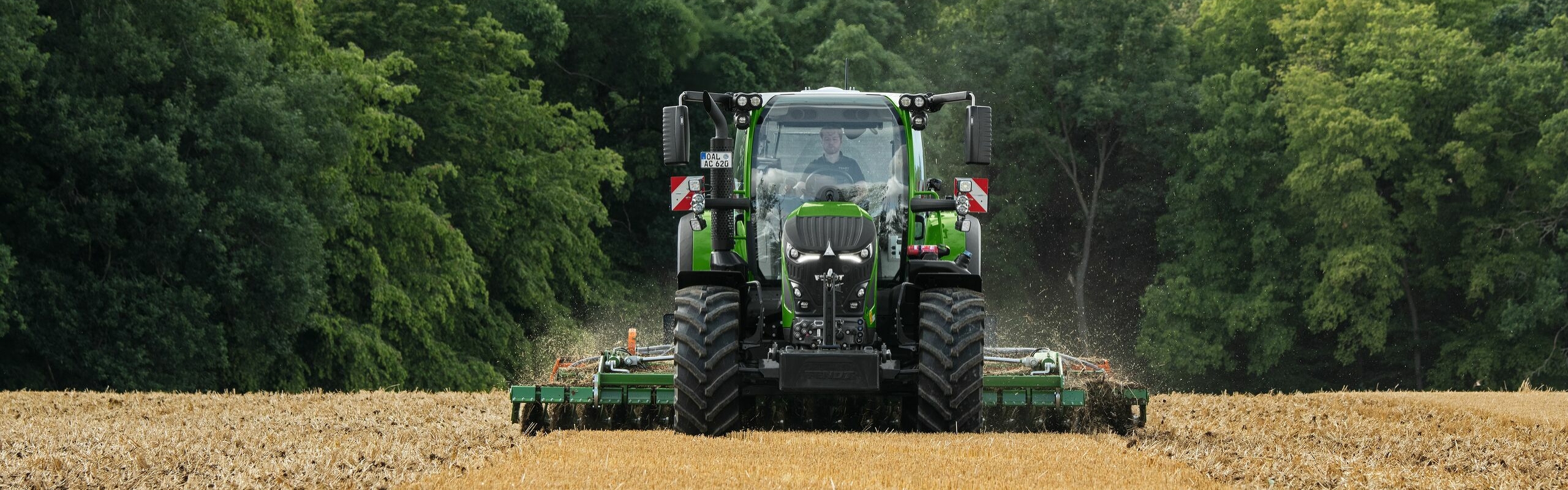 Un Fendt 600 Vario verde trabajando con un implemento en un campo marrón. Al fondo se ven un bosque en tono verde oscuro y un cielo nuboso.