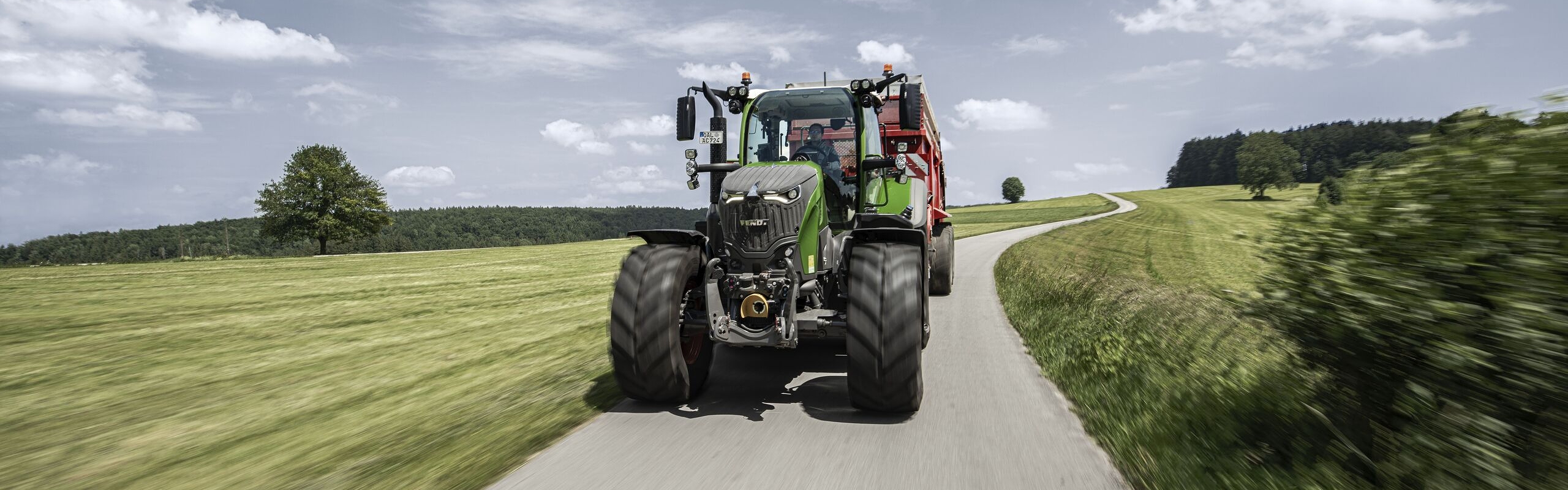 Un Fendt 700 Vario Gen7.1 circula con un implemento montado por una estrecha carretera comarcal que recorre un paisaje de montaña, bajo un cielo con algunas nubes.