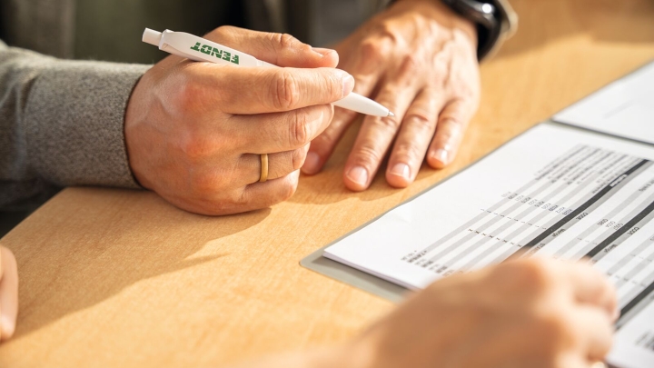 Primeros planos de dos pares de manos sobre una mesa. Una de las dos personas sostiene un bolígrafo en la mano y está firmando un documento.