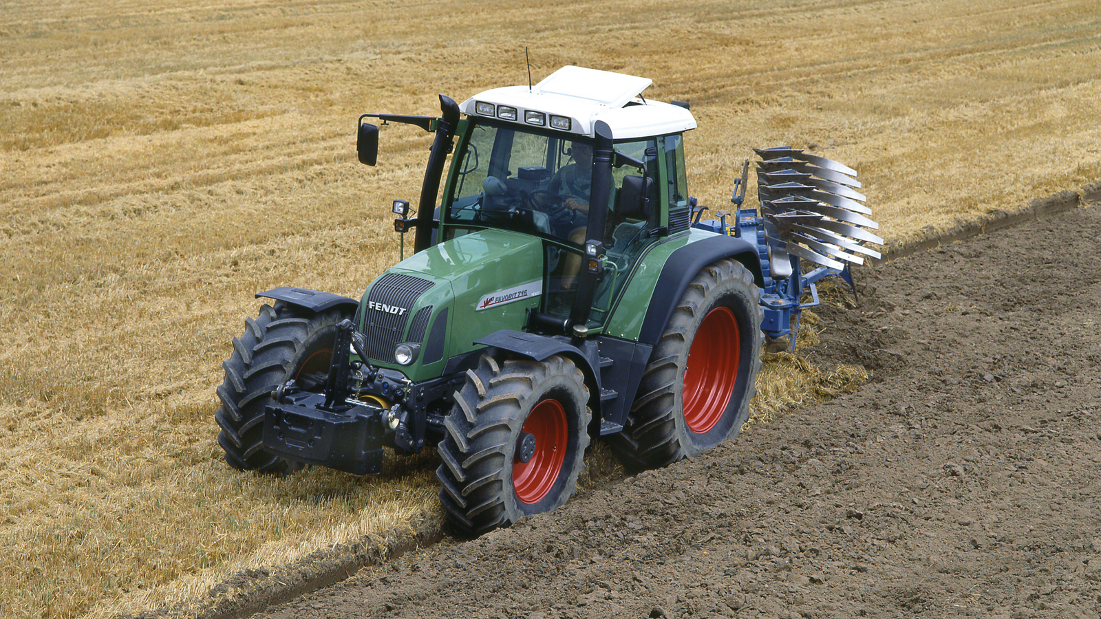 Un Fendt Favorit 700 Vario verde ara un campo que está medio cubierto de rastrojos dorados y medio recién arado.