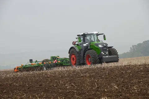 Vihreä Fendt 1000 Vario -traktori muokkaa peltoa vihreällä ja oranssilla Amazone-kultivaattorilla, hämärässä ajovalot ovat päällä.