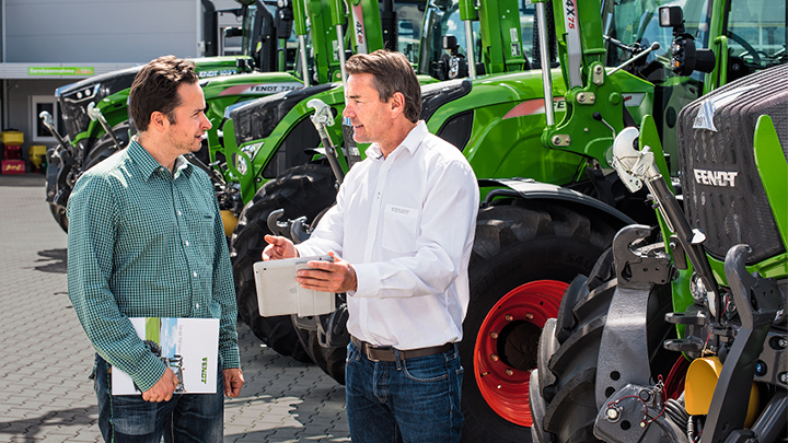 Neuvottelukeskustelu vihreiden Fendt-traktoreiden edessä Kaksi henkilöä seisoo kivetyllä pihalla ja keskustelee. Taustalla on useita vihreitä Fendt-traktoreita, joissa on suuret mustat renkaat ja punaiset vanteet, rivissä. Toinen henkilö pitää kiinni leikekirjasta, toinen on pukeutunut ruudulliseen paitaan ja p