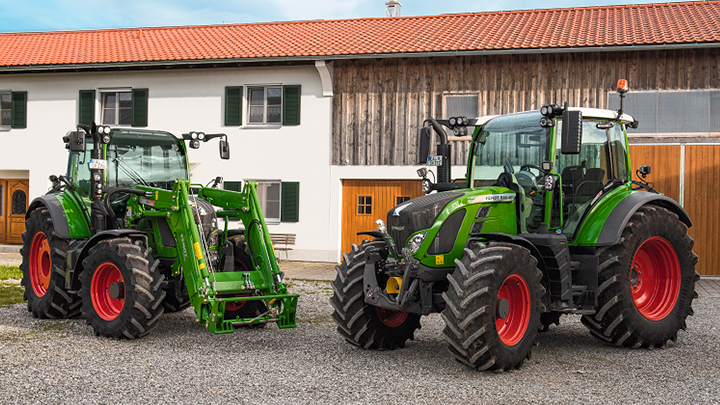Kaksi vihreää Fendt-traktoria punakattoisen maatilan edessä Kaksi vihreää Fendt-traktoria, joissa on suuret mustat renkaat ja punaiset vanteet, seisoo sorapihalla maatilan edessä. Maatilan julkisivu on puuta ja valkoista kipsiä, ikkunaluukut ovat vihreät ja katto on punatiilinen. Toisessa traktorissa on etukuormaa
