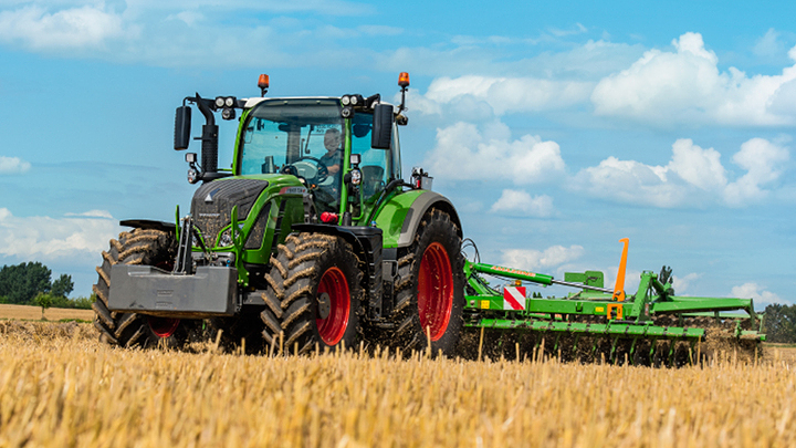 Fendt-traktori työskentelee viljapellolla sinisen taivaan alla Vihreä Fendt-traktori, jossa on suuret mustat renkaat ja punaiset vanteet, ajaa kultaisen peltoon. Takana on vihreä maatalouskone. Taustalla on sininen taivas ja valkoisia pilviä.