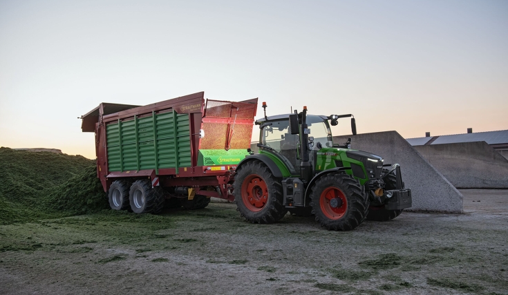 Fendt 500 Vario purkaa Strautmann-noukinvaunua aumalla Vihreä Fendt 500 Vario purkaa vihreä-punaista Strautmann-noukinvaunua aumalla hämärässä.