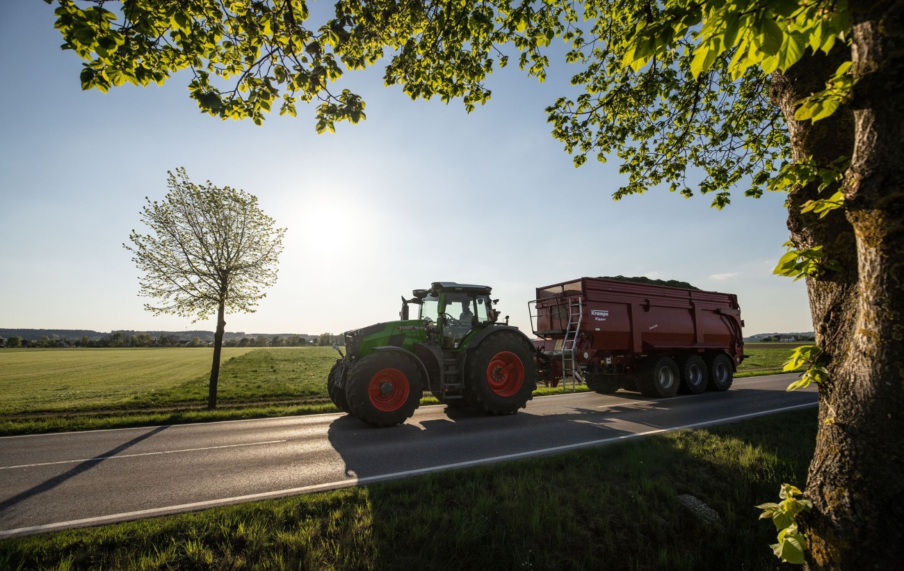 Fendt 800 Vario maantiekuljetuksessa täyden perävaunun kanssa Vihreä Fendt 800 Vario ajaa täyteen lastatun perävaunun kanssa tietä pitkin, kuva on kehystetty oikealta puolelta puulla ja traktori heittää pitkät varjot.