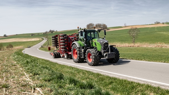Fendt 700 Vario Gen7 noukinvaunun kanssa peltojen välisellä tiellä. Fendt 700 Vario Gen7.1 ja siihen kiinnitetty työkone ajavat maantietä mäkisessä kevätmaisemassa.